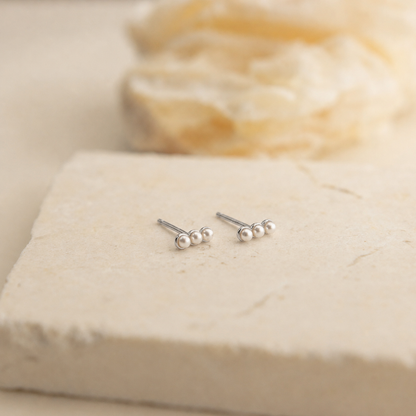 A pair of small silver stud earrings with three tiny white pearl-like beads on each earring, displayed on a light beige stone slab with a soft blurred cream background.