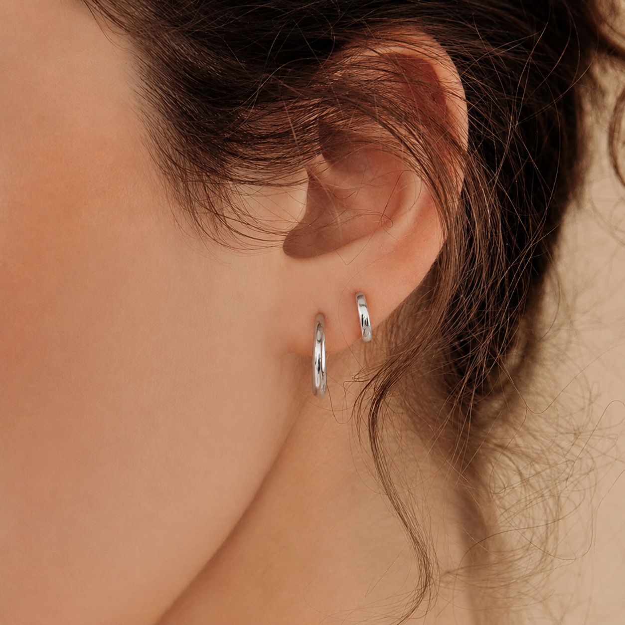 Close-up of a person wearing two silver hoop earrings with a neutral background