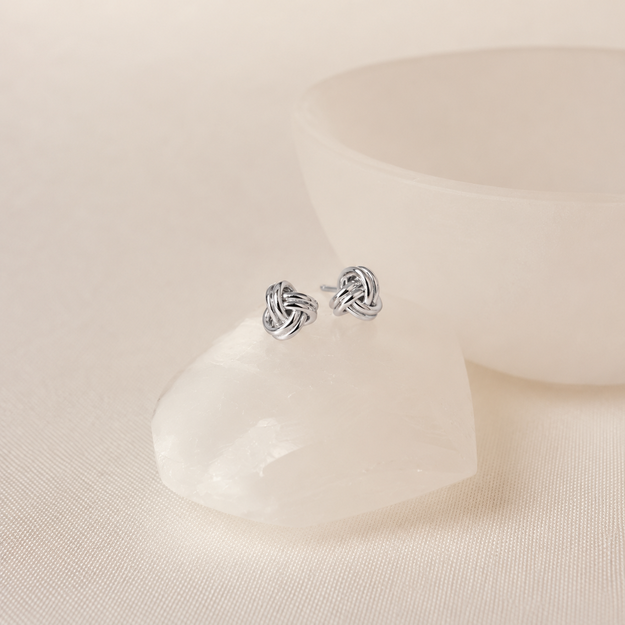 A pair of silver stud earrings with a knotted ribbon design, displayed on a clear crystal cluster with a soft-focus white ceramic bowl in the background.