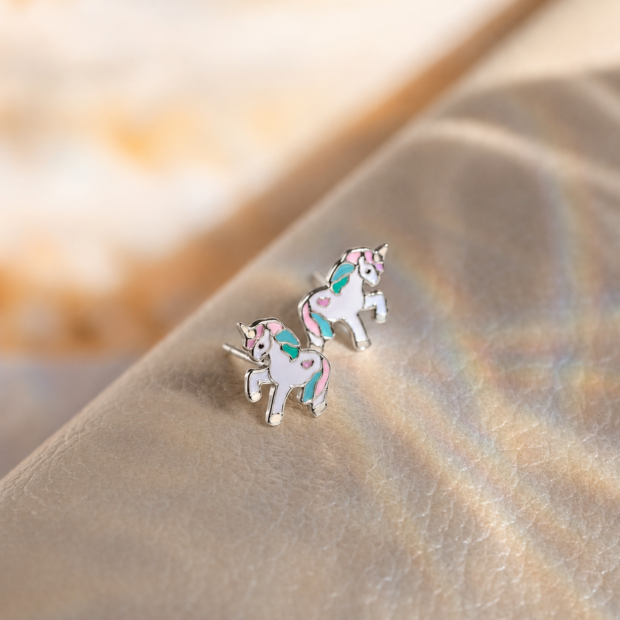 A pair of silver unicorn stud earrings with pastel pink, teal, and white enamel details, resting on a textured beige surface with a soft-focus background.