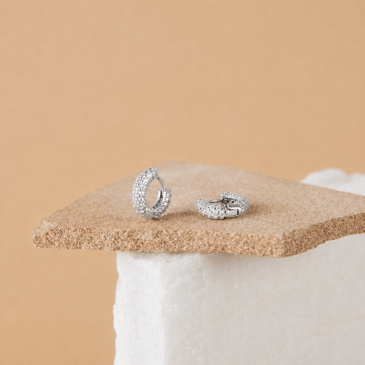 A pair of silver-toned hoop earrings covered in small clear stones, displayed on a textured tan stone slab against a warm beige background.