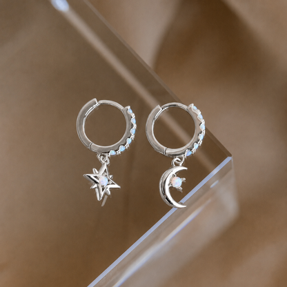 Silver hoop earrings with star and moon charms on a reflective surface.