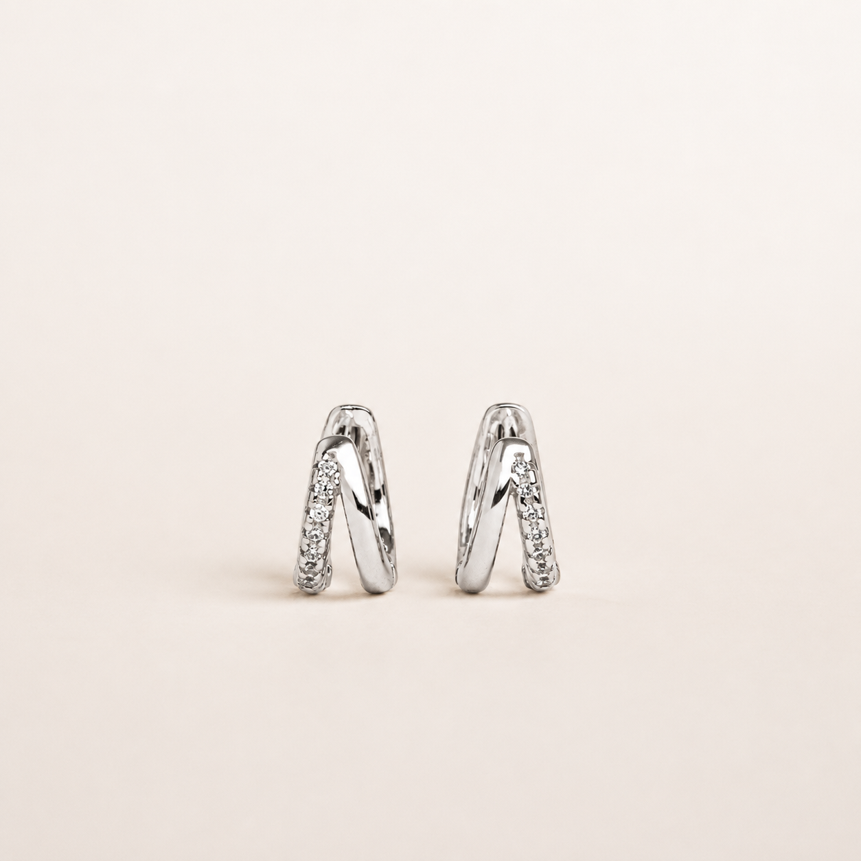 A pair of silver-toned huggie hoop earrings with pavé-set clear stones, shown on a plain light beige background.
