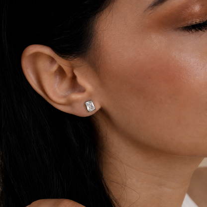 A close-up of a model's ear wearing a small rectangular silver stud earring with a clear gemstone.