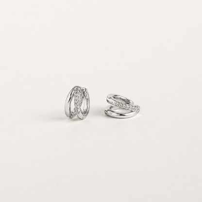 A pair of silver-toned huggie hoop earrings with a twisted, layered design, one earring featuring a row of small clear stones, photographed on a clean, light off-white background.