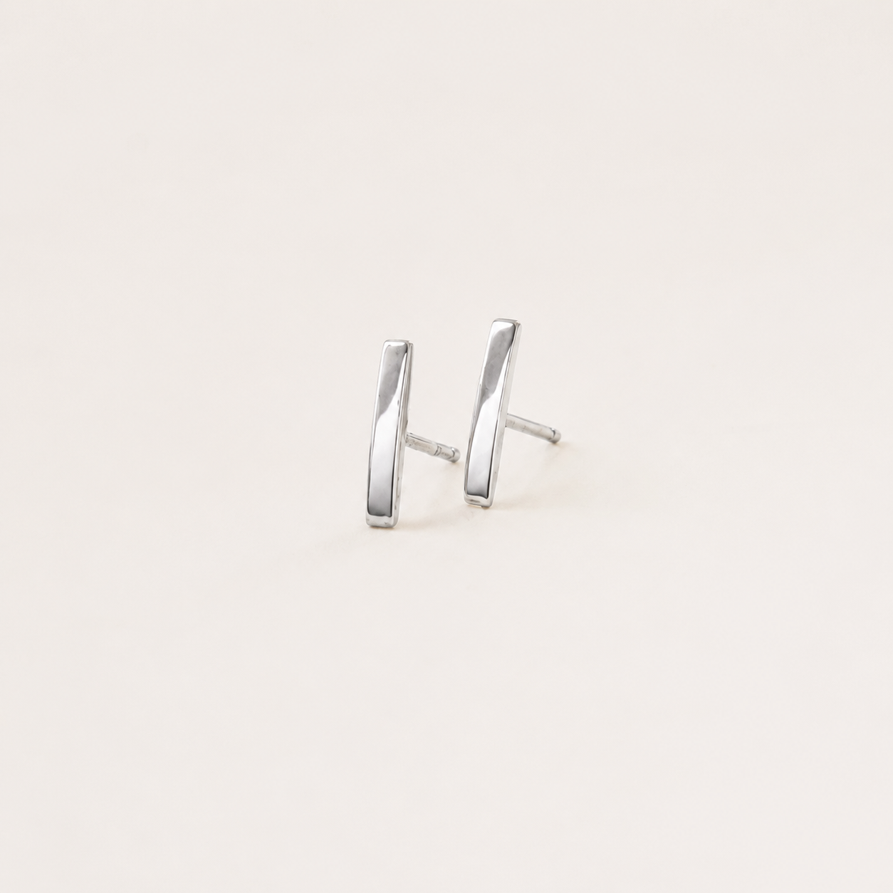 A pair of minimalist silver stud earrings with a modern, slightly angled bar design, shown on a plain off-white background.