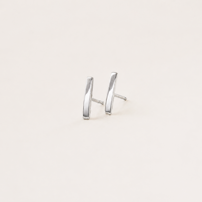 A pair of minimalist silver stud earrings with a modern, slightly angled bar design, shown on a plain off-white background.
