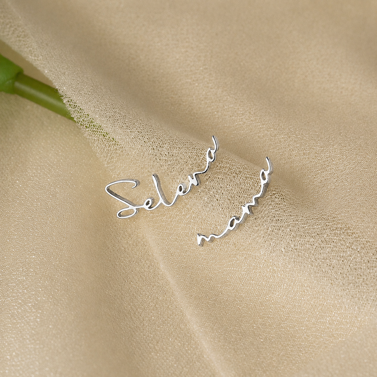 Silver earrings with 'Selena' and 'Mom' engraved on a beige fabric background