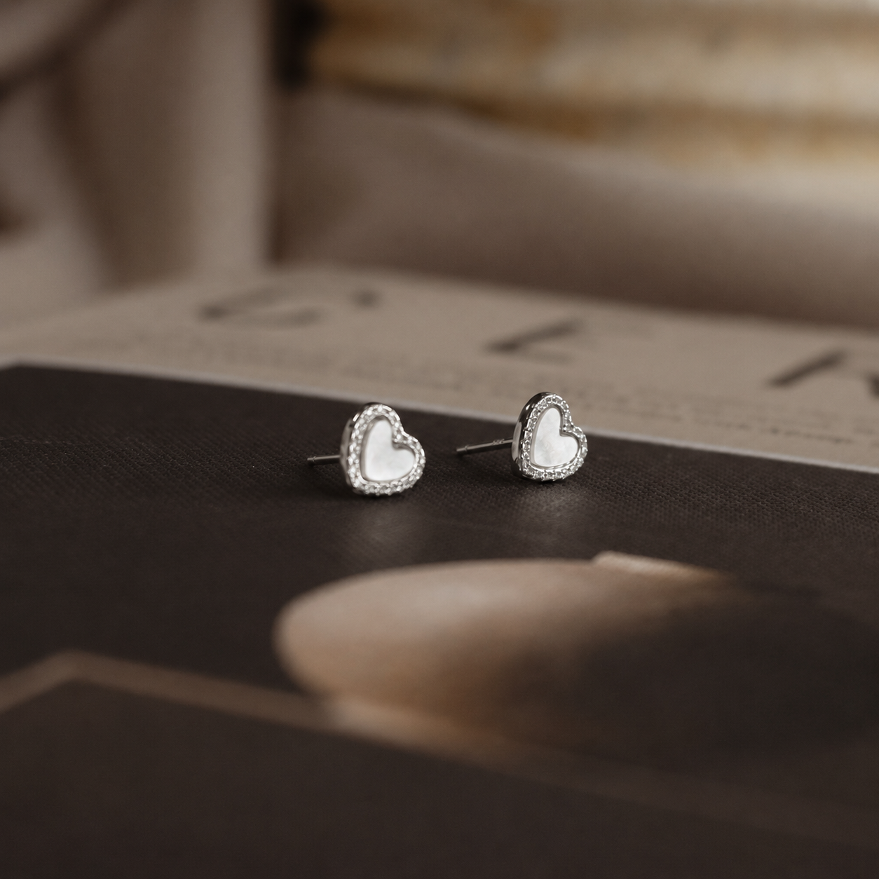A pair of silver heart-shaped stud earrings with white inlay and a delicate rhinestone border, resting on a dark textured surface.