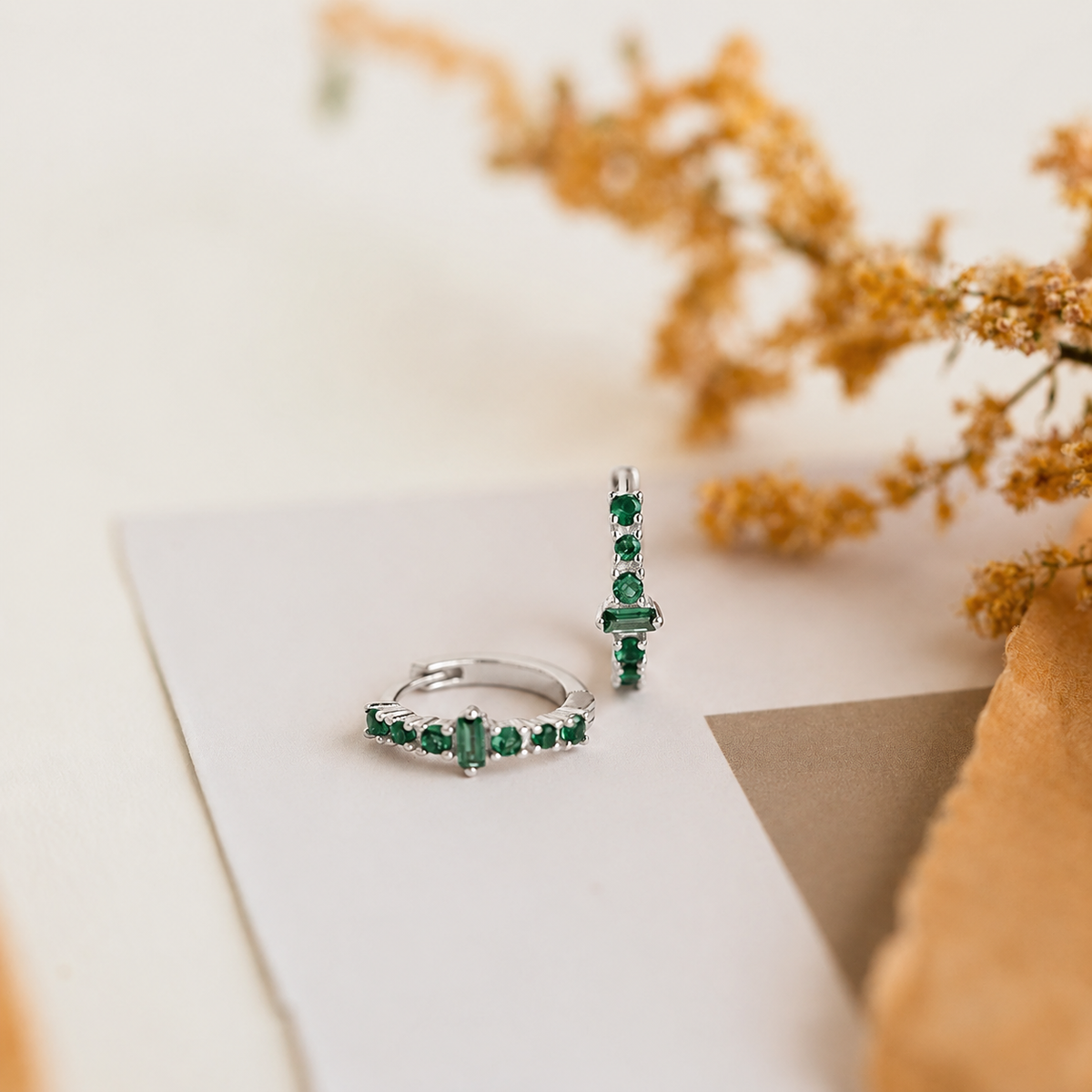 Pair of emerald green gemstone rings on a white surface with dried flowers in the background