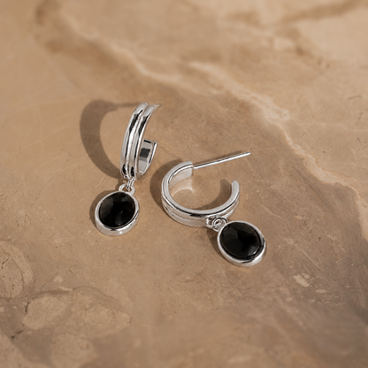 Silver hoop earrings with black stones on a beige stone surface