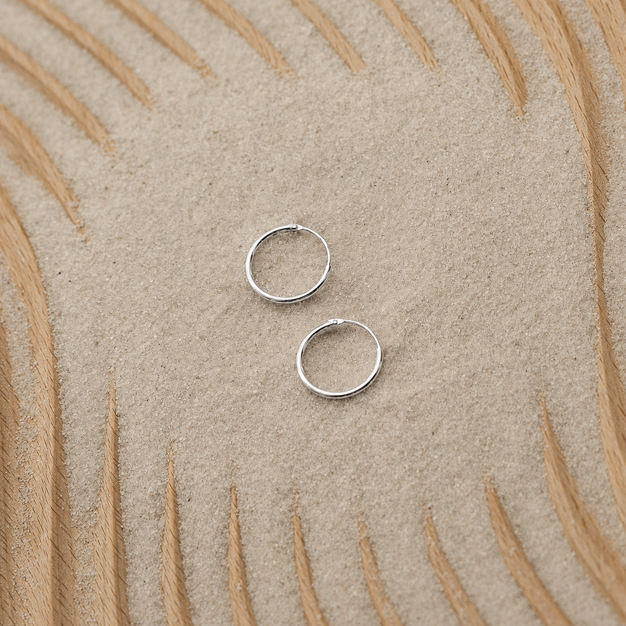 Two small silver hoop earrings placed on a textured beige surface with a curved tan pattern in the background.