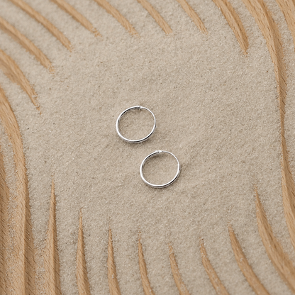 Two small silver hoop earrings placed on a textured beige surface with a curved tan pattern in the background.