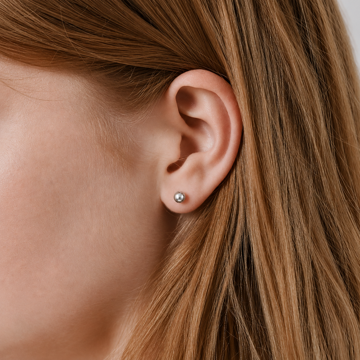 A close-up of a person's ear wearing a small round silver stud earring, with brown hair framing the side of the face.