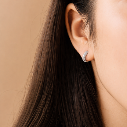 A close-up of a woman's ear wearing a small crescent-shaped stud earring set with clear stones, against a warm beige background.