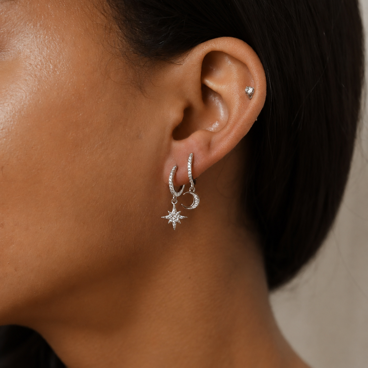 A close-up of a model's ear wearing a silver huggie hoop earring with a dangling crescent moon and star charm, alongside a small stud earring in the lobe.