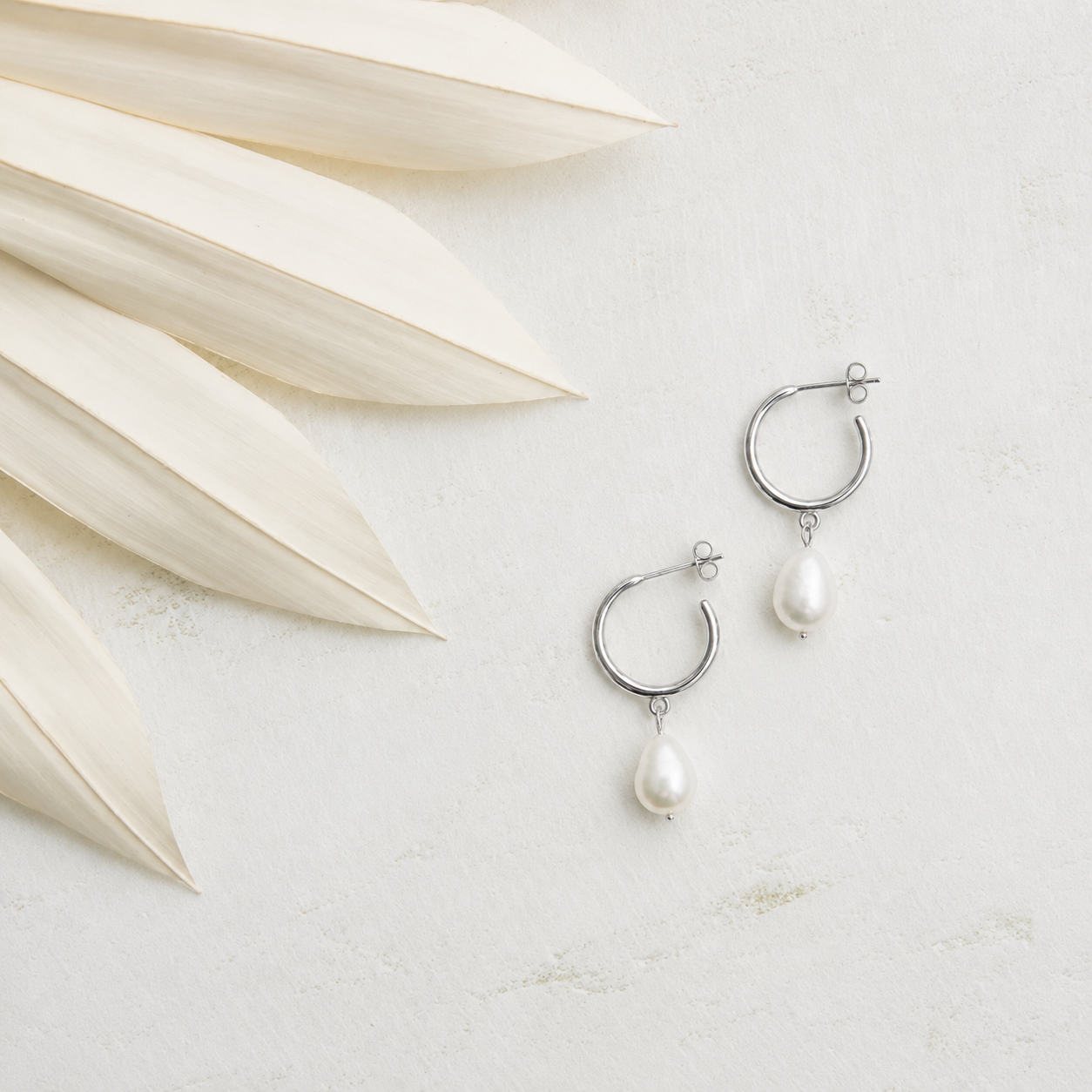 Silver hoop earrings with pearls on a light surface with dried leaves.