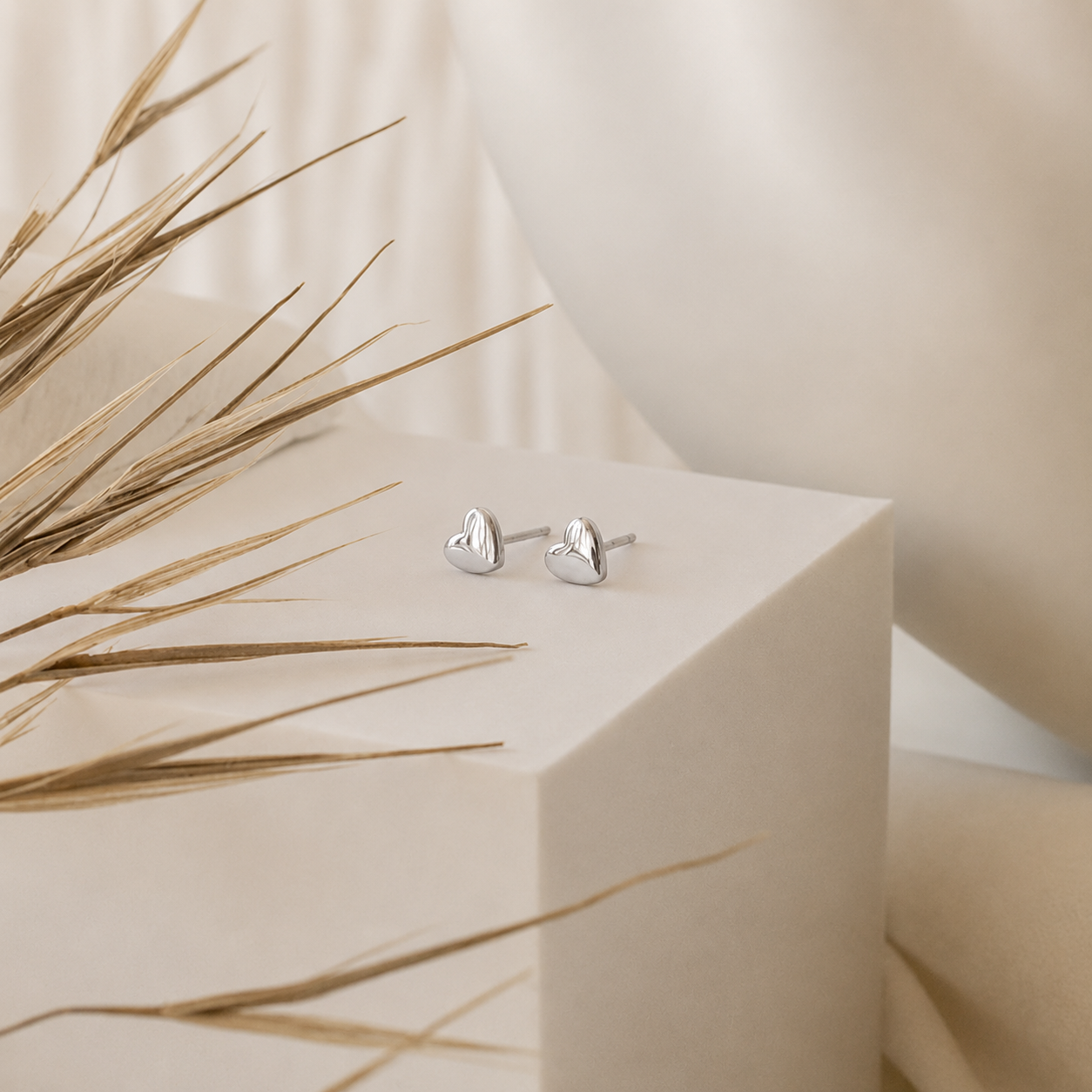 Silver heart-shaped earrings on a beige cube with dried grasses.