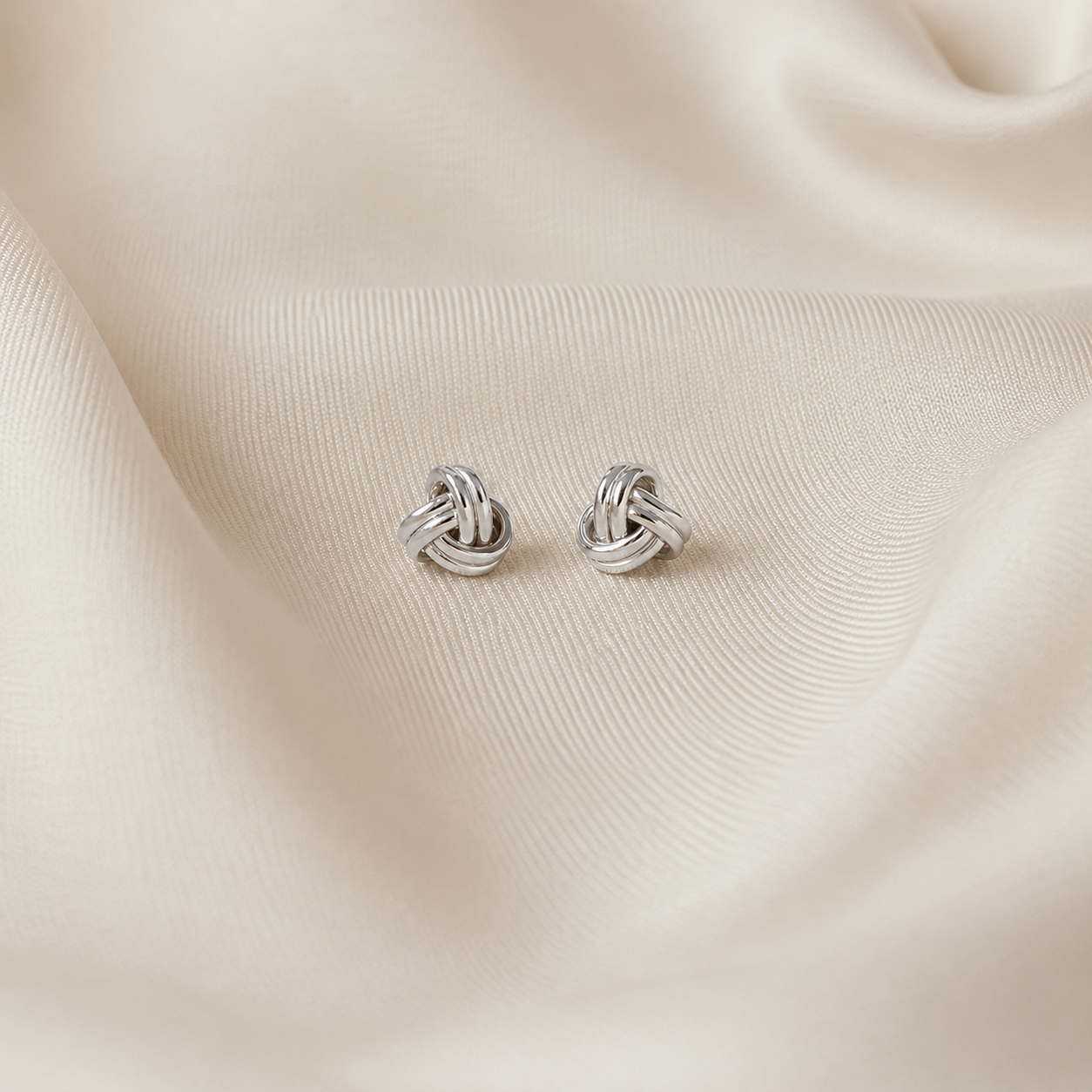 A pair of small silver knot stud earrings resting on draped cream-colored satin fabric.