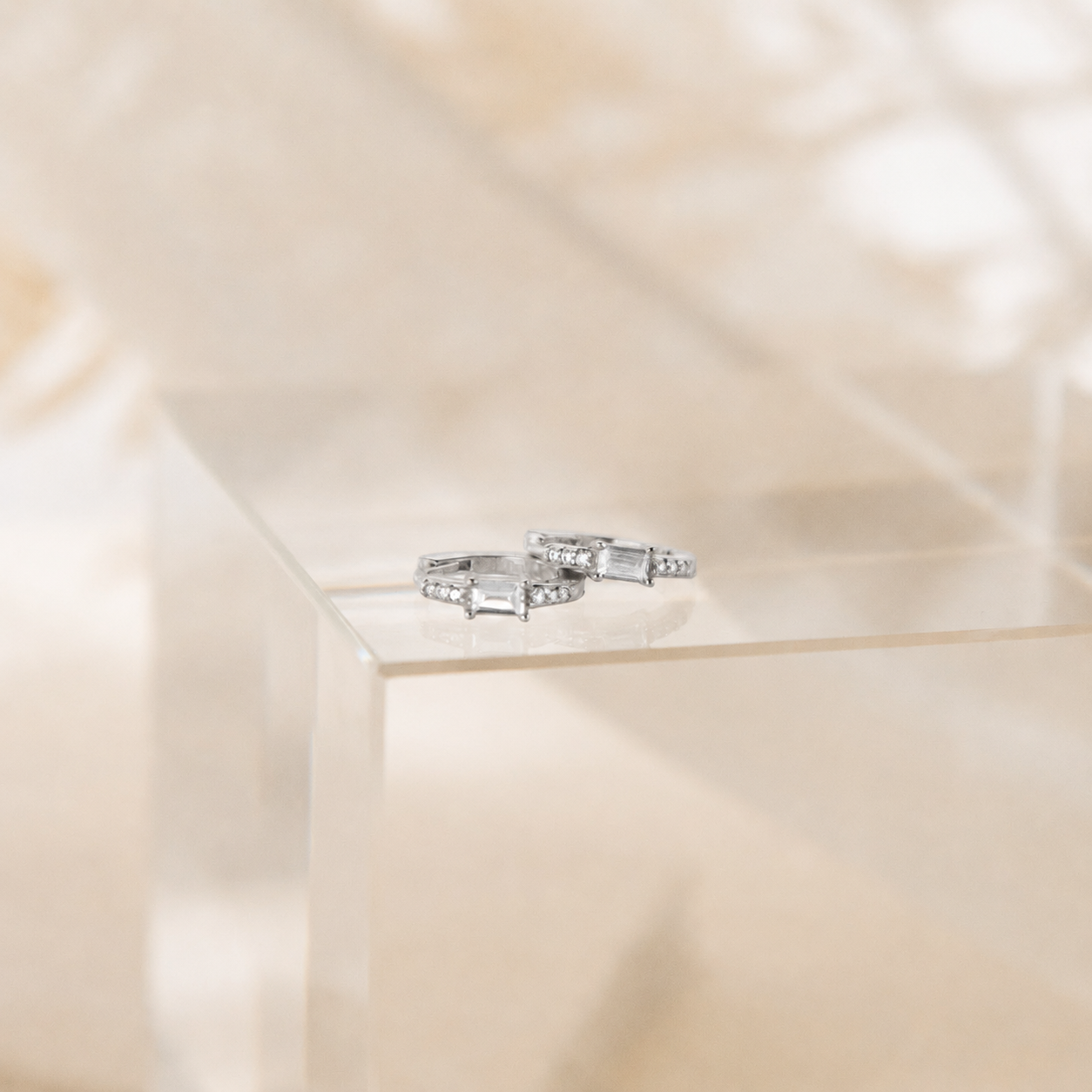 A pair of silver rings with rectangular baguette-cut clear stones, displayed on a clear acrylic block against a soft beige background.