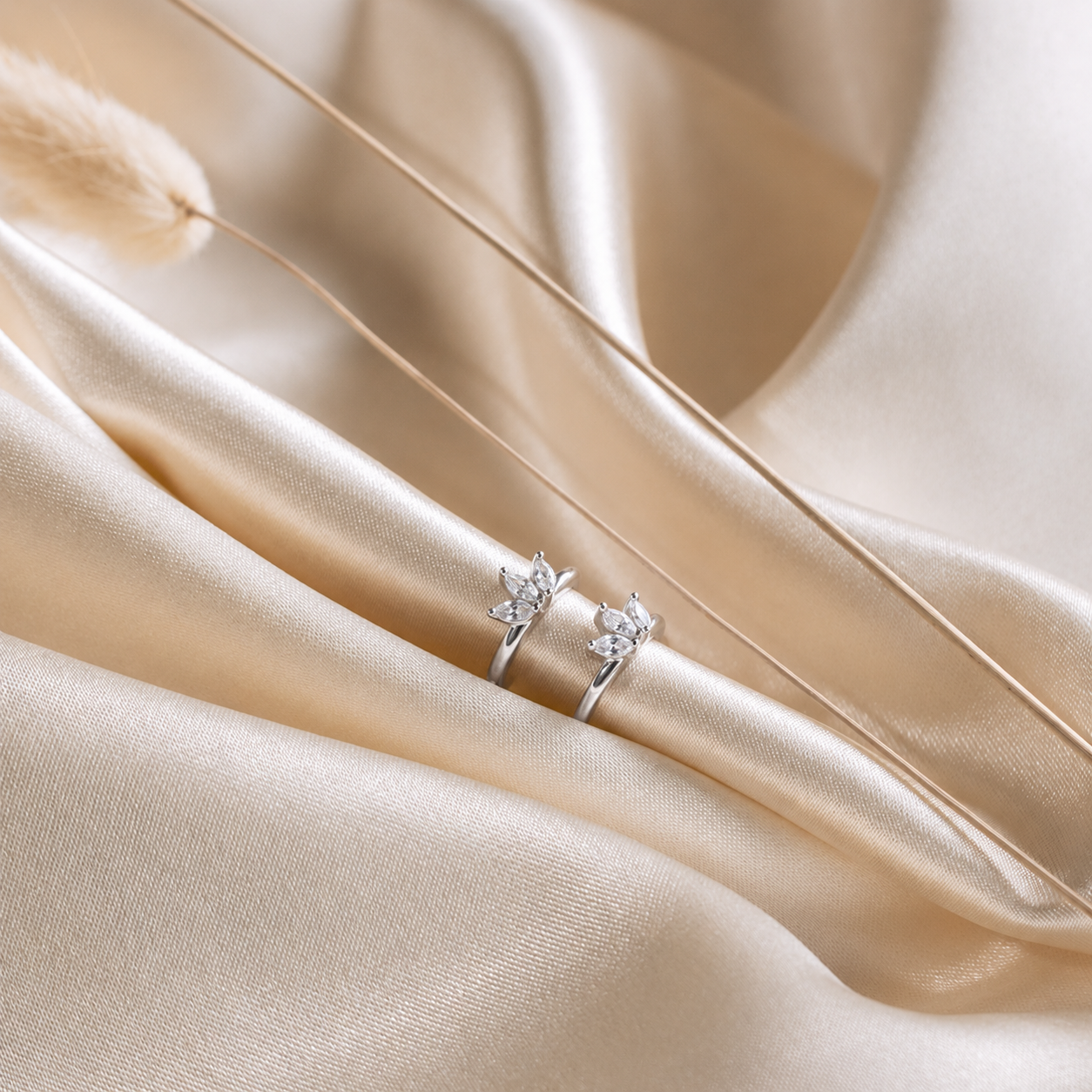 A pair of silver rings with small floral or leaf-shaped clear stones, displayed on draped champagne satin fabric.