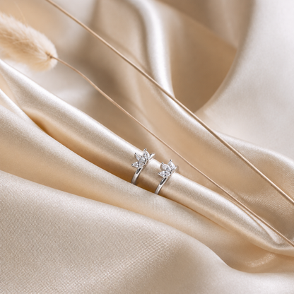 A pair of silver rings with small floral or leaf-shaped clear stones, displayed on draped champagne satin fabric.