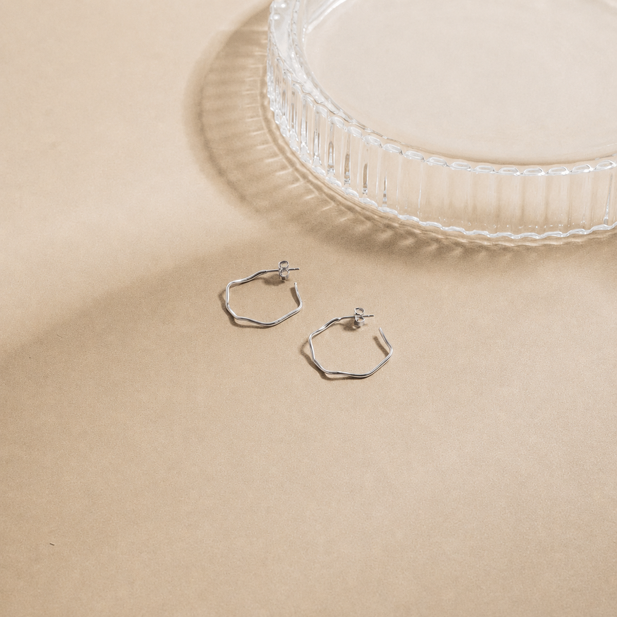 Silver hoop earrings on a beige surface with a clear glass dish in the background
