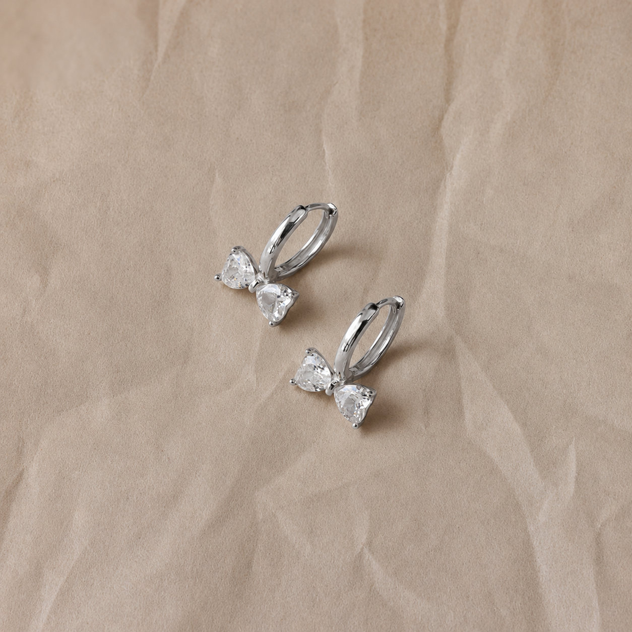 Silver bow-shaped earrings with clear gemstones on a beige fabric background