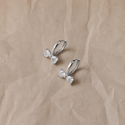Silver bow-shaped earrings with clear gemstones on a beige fabric background