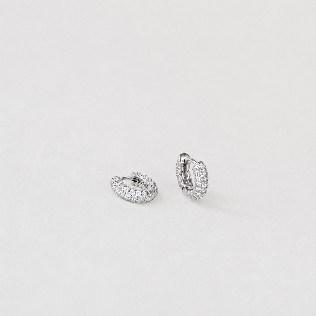 A pair of small silver huggie hoop earrings paved with clear stones, displayed on a clean, light gray background.