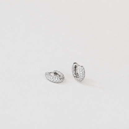 A pair of small silver huggie hoop earrings paved with clear stones, displayed on a clean, light gray background.