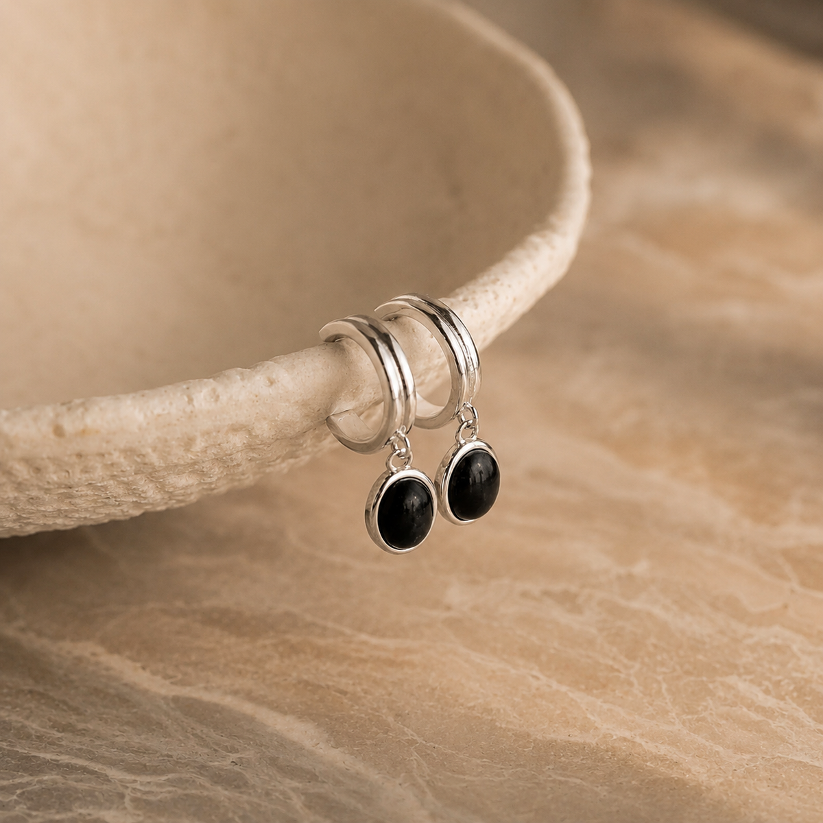 Silver hoop earrings with black stones on a beige stone surface