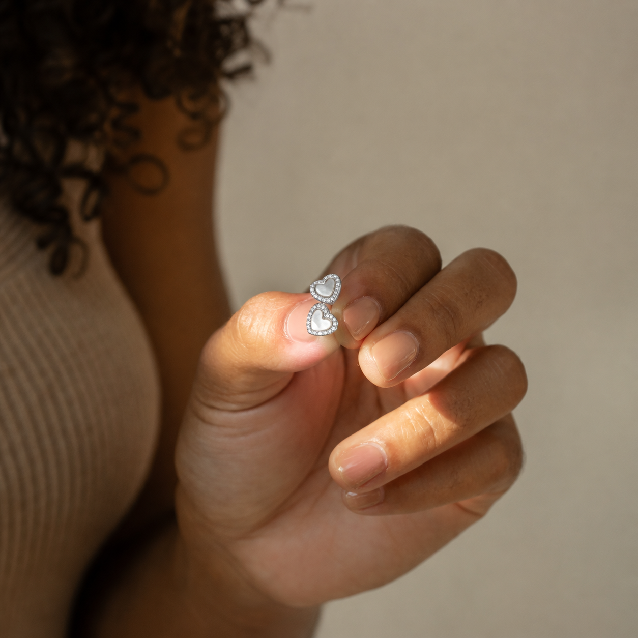 A close-up of a hand holding a pair of silver double-heart stud earrings with clear stones, set against a soft beige background.