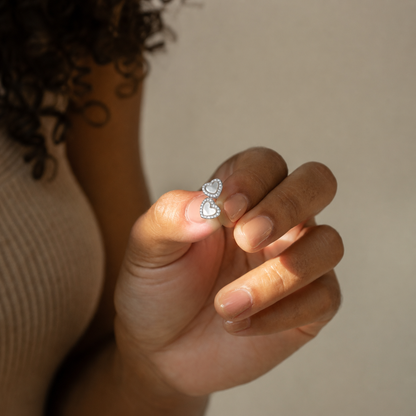 A close-up of a hand holding a pair of silver double-heart stud earrings with clear stones, set against a soft beige background.