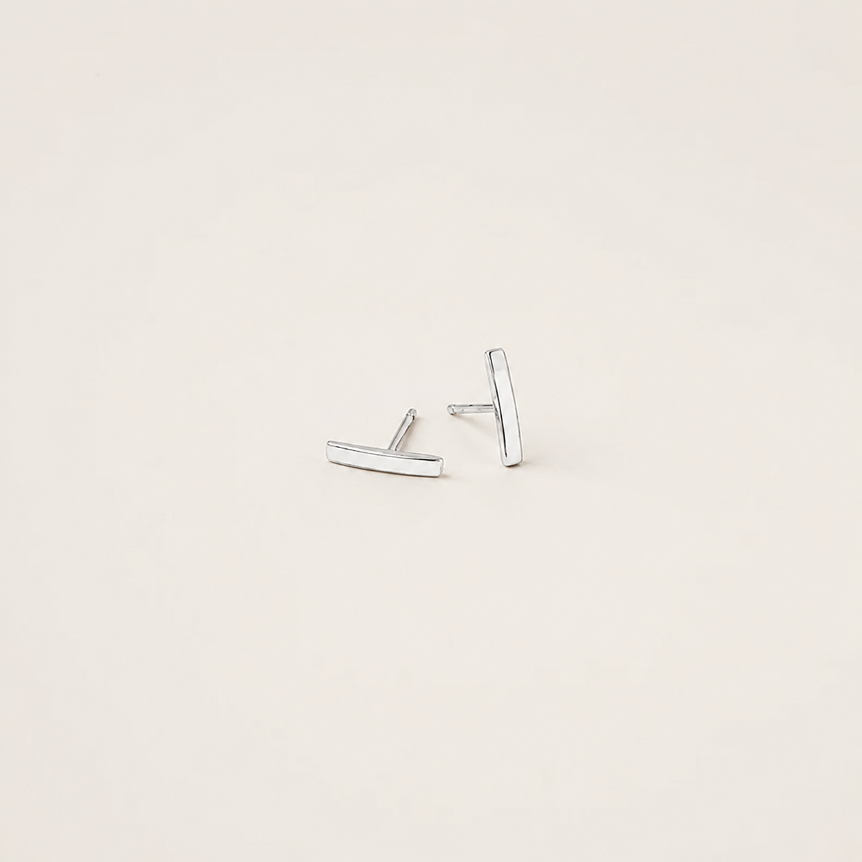 A pair of minimalist silver bar stud earrings on a warm off-white background.