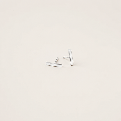 A pair of minimalist silver bar stud earrings on a warm off-white background.