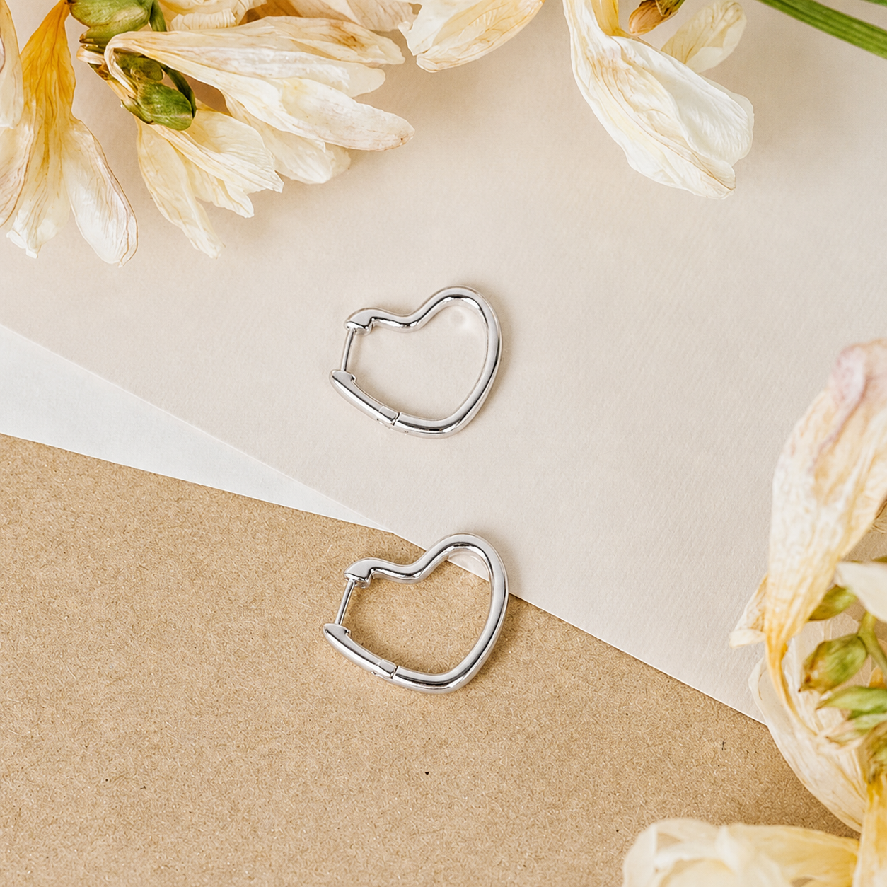 Heart-shaped silver hoop earrings on a beige and white paper background with flowers.