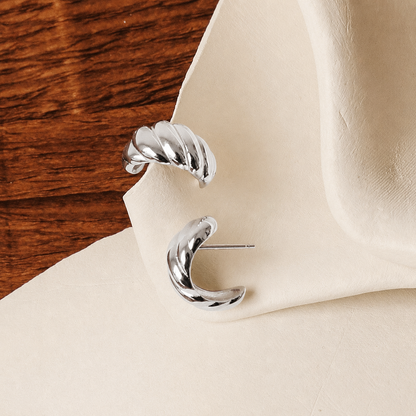 A pair of polished silver crescent-shaped hoop earrings displayed on a cream-colored textured surface with a dark wood background.