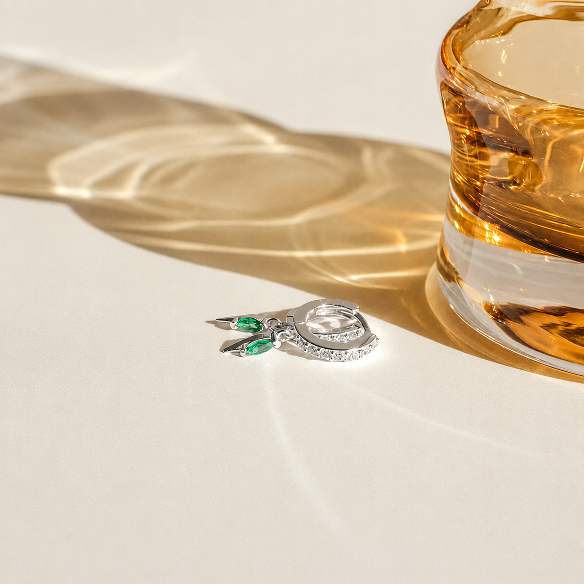 Two silver earrings with green gemstones on a white surface next to a glass of whiskey.