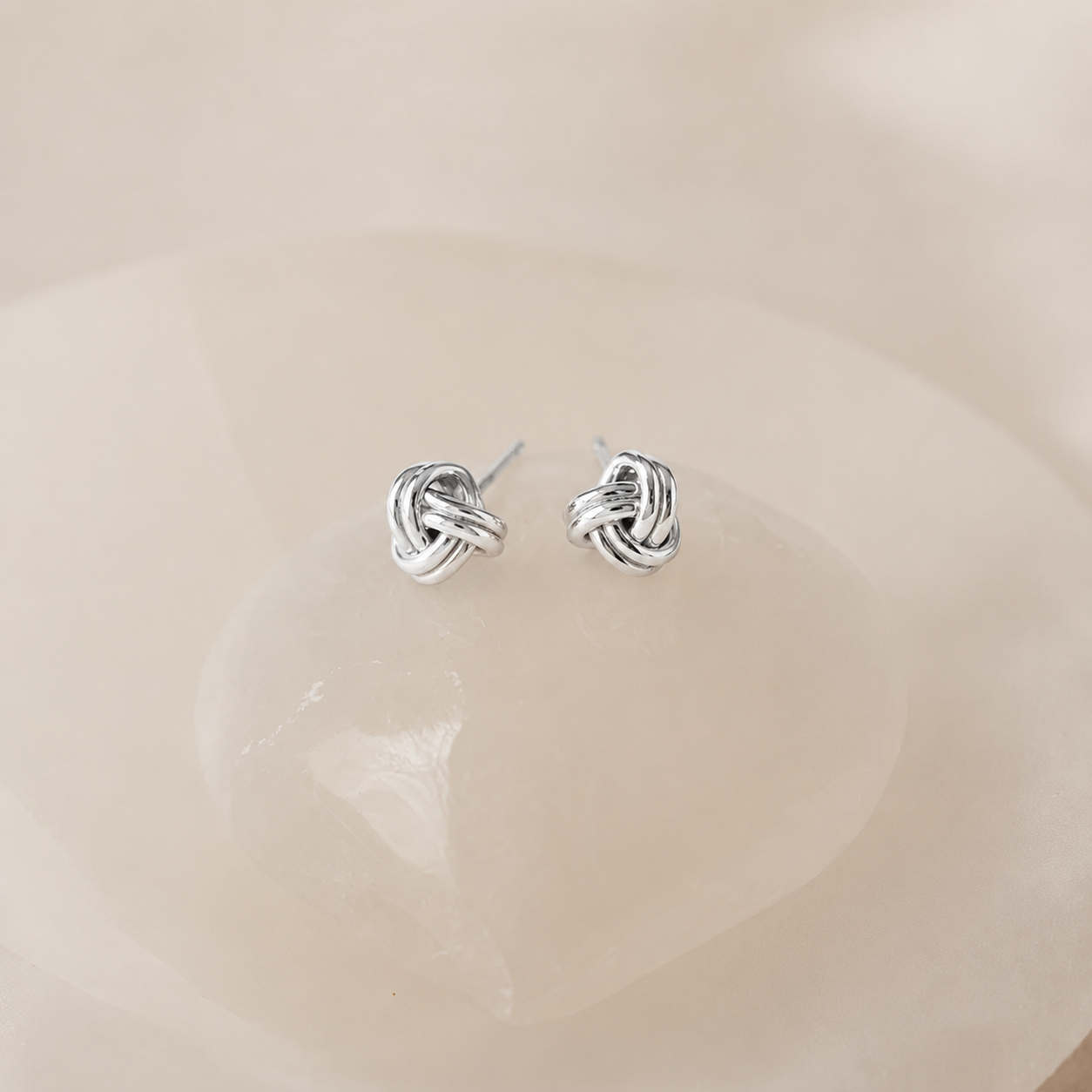 A pair of small silver knot stud earrings resting on a smooth, pale pink stone dish.