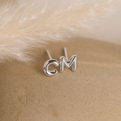 Silver 'CM' letter earrings on a textured beige surface with white fur background